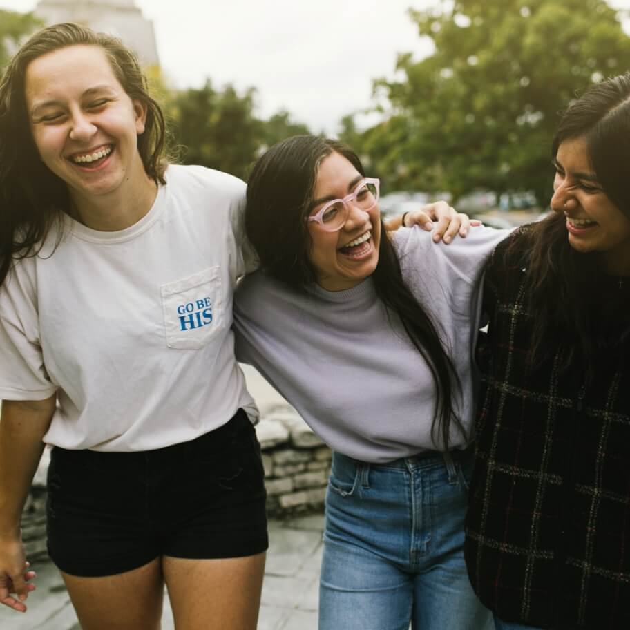 Three girls have their arms around each other laughing on a college campus where they work as missionaries providing Catholic evangelism.