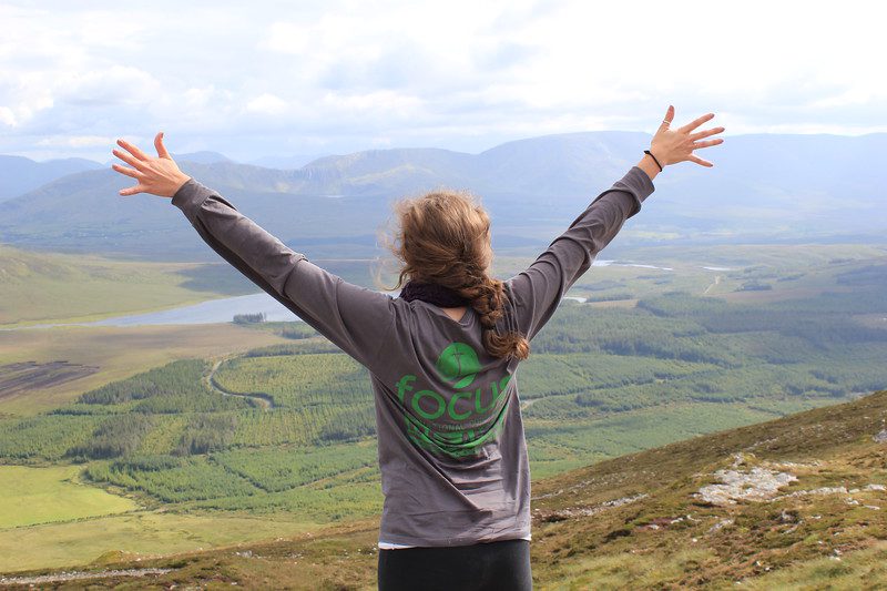 A student on a FOCUS mission trip, wearing a FOCUS shirt lifting her arms up to the sky looking over a green landscape with mountains. 