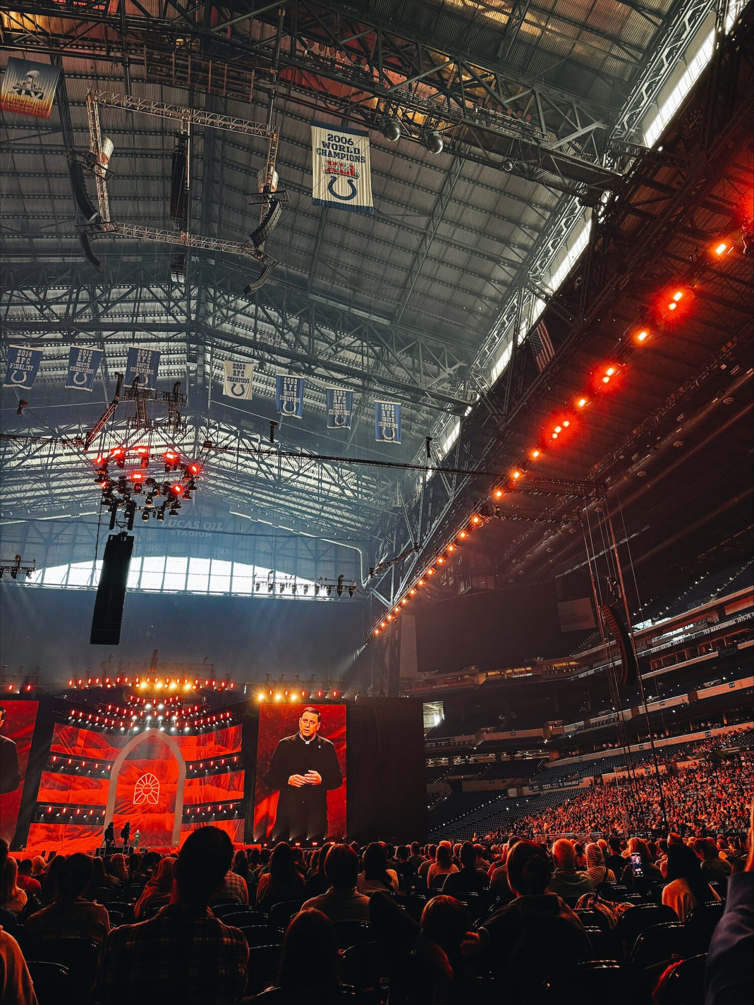 The image shows a full stadium and a priest speaking on the stage at the National Eucharistic Congress