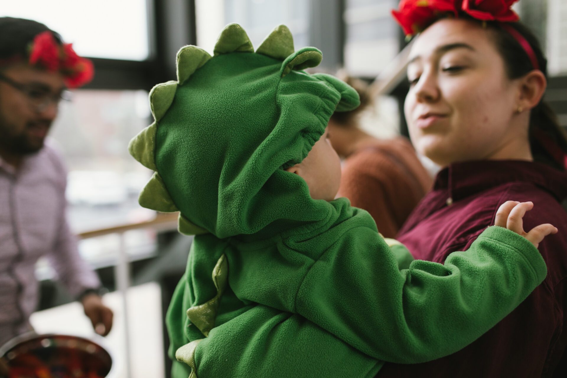 Wondering "can Catholics celebrate Halloween?" This post looks at what the answer is. The image shows a baby in a dinosaur costume and his mother holding him in a flower crown.