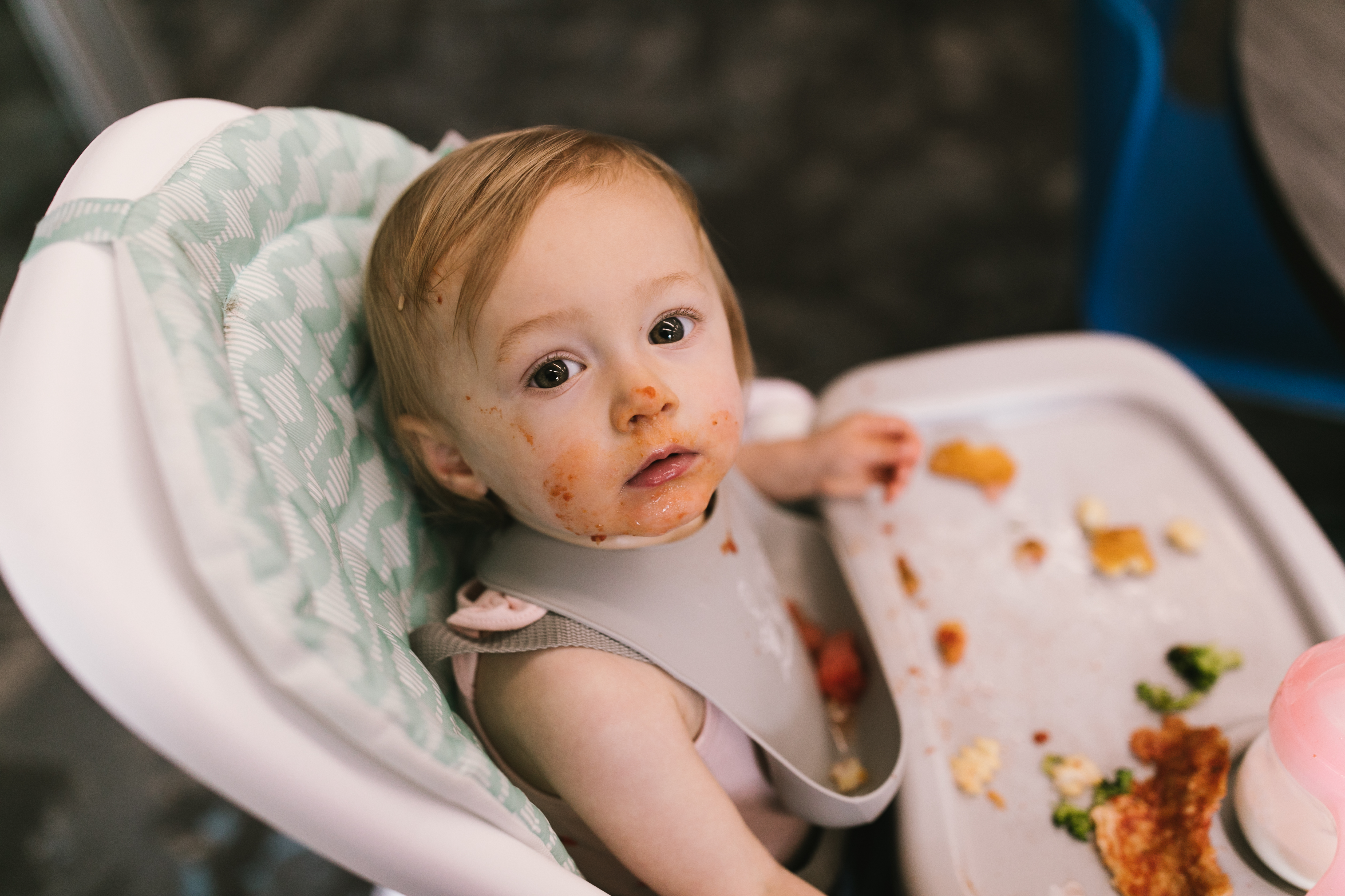 A baby looks up to the camera with food on her face and hands after eating a saint-themed meal. 